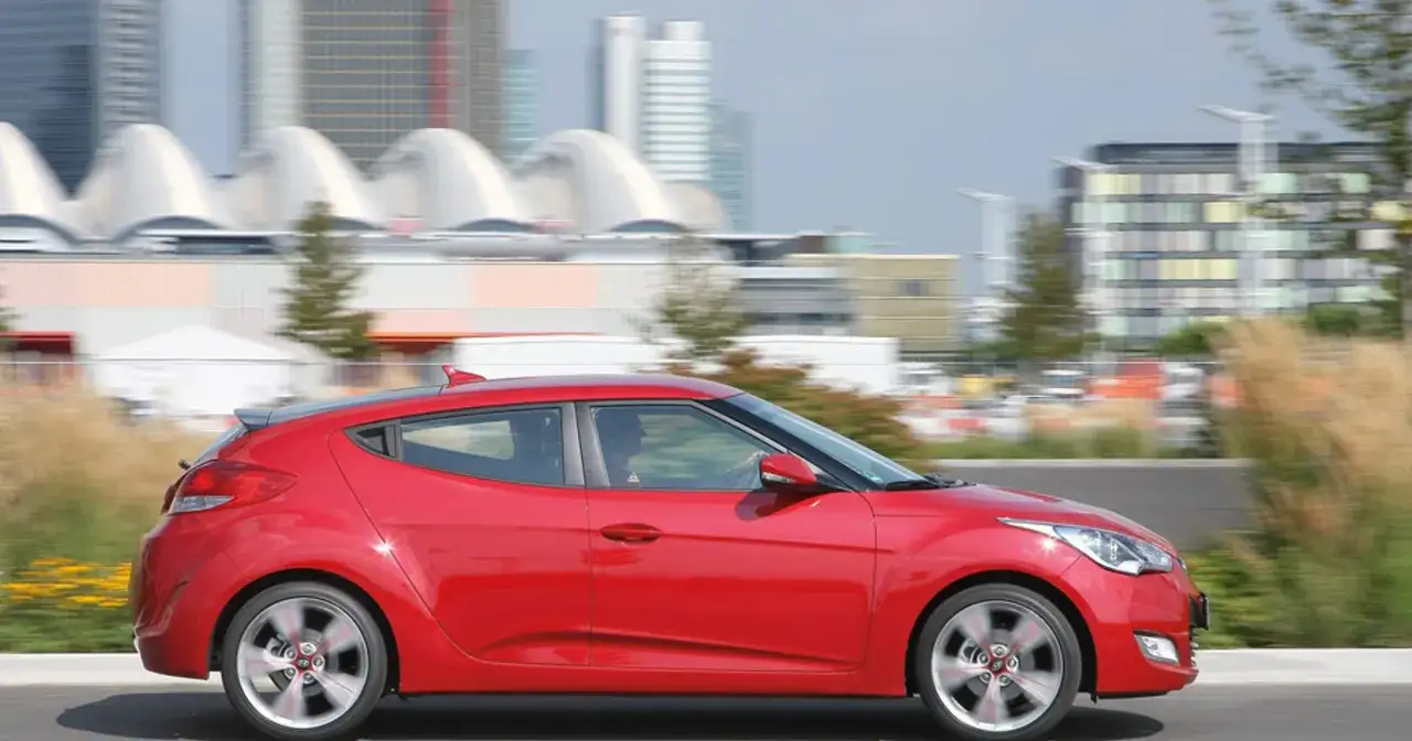 Czerwony Hyundai Veloster w ruchu miejskim, z rozmytym tłem budynków i zieleni.
