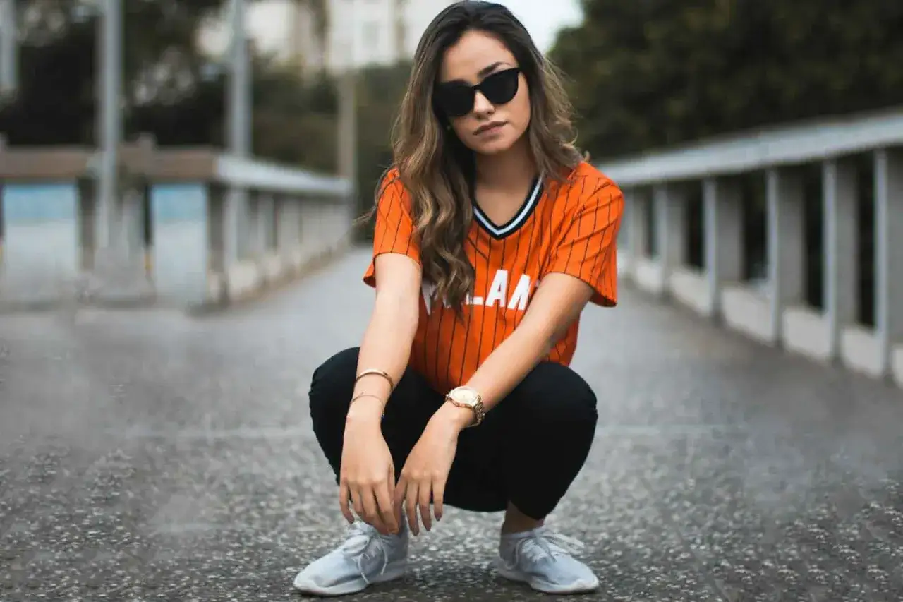 Joven con gafas de sol y camiseta naranja a rayas, pantalones negros y zapatillas deportivas, mostrando diferentes estilos de ropa urbanos.