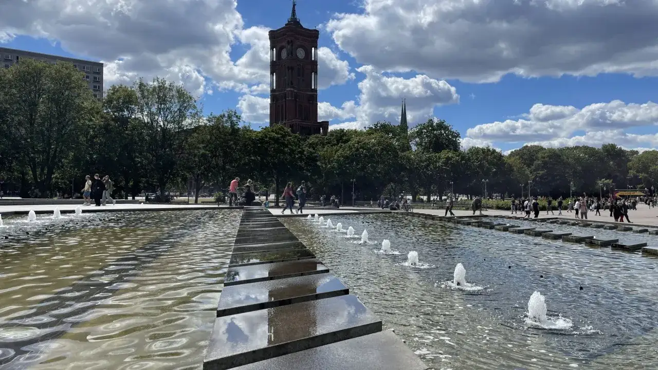 Brunnenanlage am Alexanderplatz Berlin mit dem Rathausturm im Hintergrund. Menschen spazieren und genie&szlig;en den sonnigen Tag.