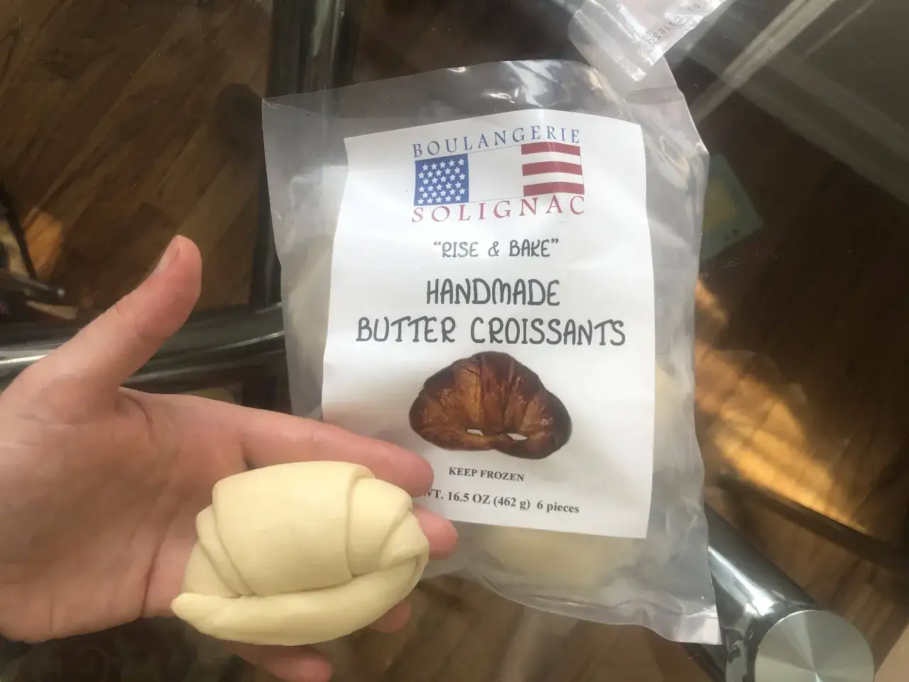 Mano sosteniendo un croissant crudo junto a un paquete de croissants de mantequilla hechos a mano. ¡Se pueden congelar los croissants para hornearlos más tarde!