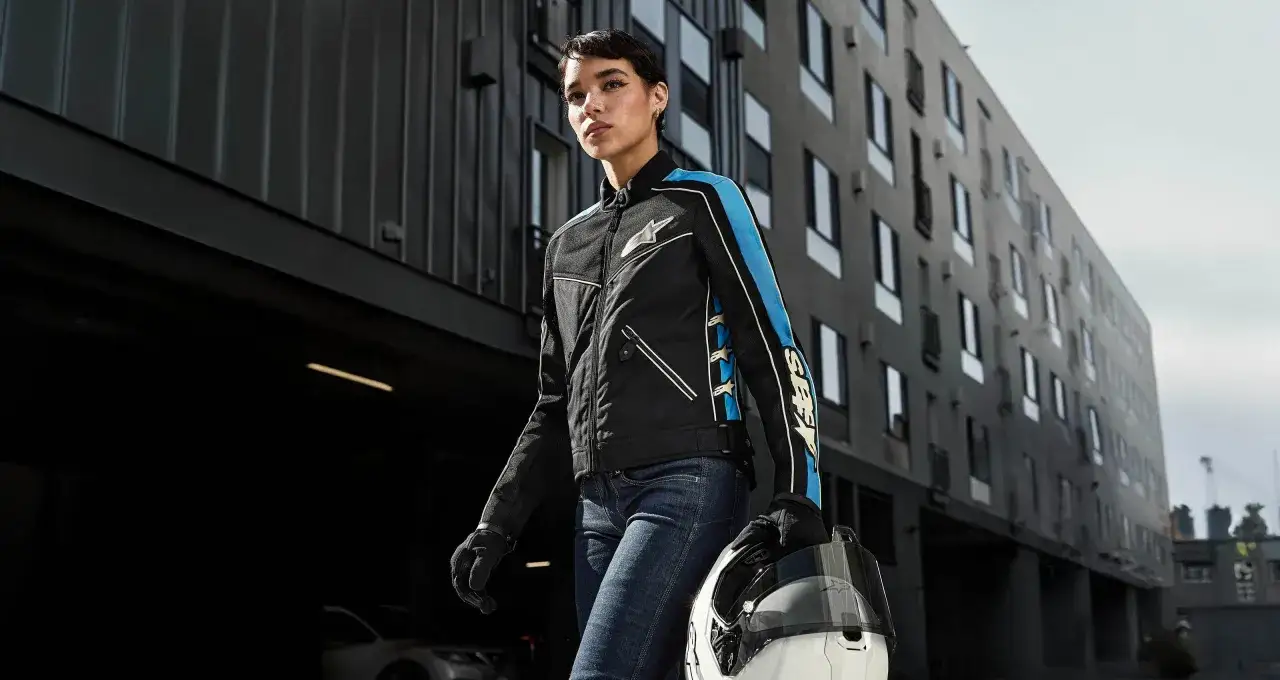 Kobieta w kurtce Alpinestars Chrome Sport z niebieskimi akcentami i gwiazdami, trzymająca biały kask, idzie obok nowoczesnego budynku.