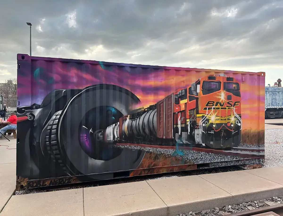 Un tren BNSF emerge da un obiettivo di fotocamera in un murale su un container, un'opera che ricorda lo stile di Mike Brodie.