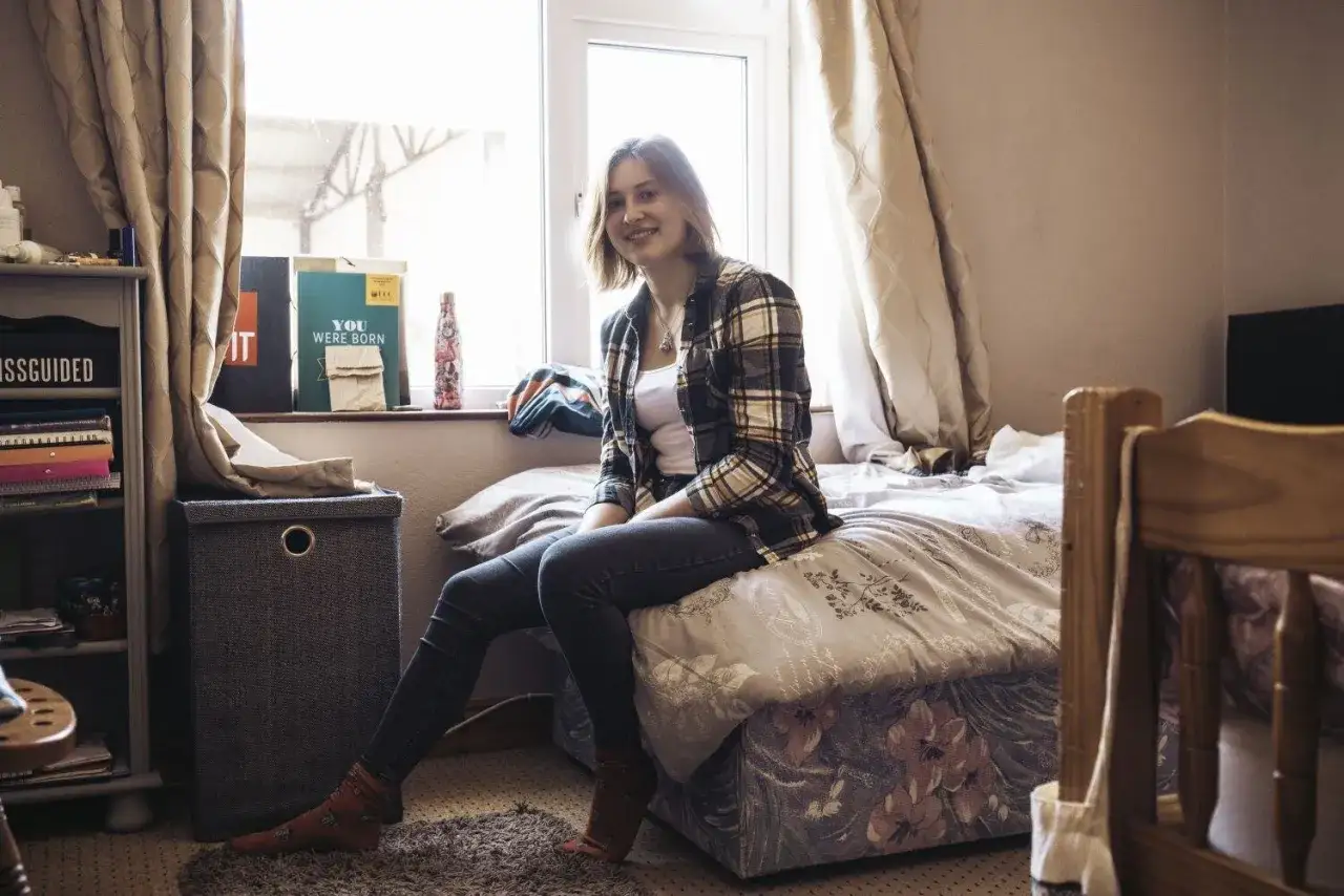 Junge Frau sitzt l&auml;chelnd auf ihrem Bett in einem Zimmer in England. Ihr Auslandsjahr beginnt.