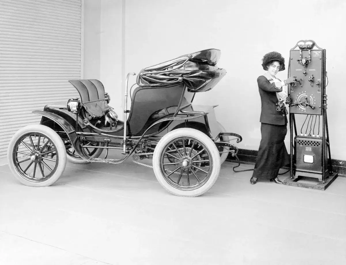 Una mujer ajusta el panel de control de lo que parece ser el primer coche el&eacute;ctrico de la historia, un veh&iacute;culo de &eacute;poca con ruedas de radios y capota.