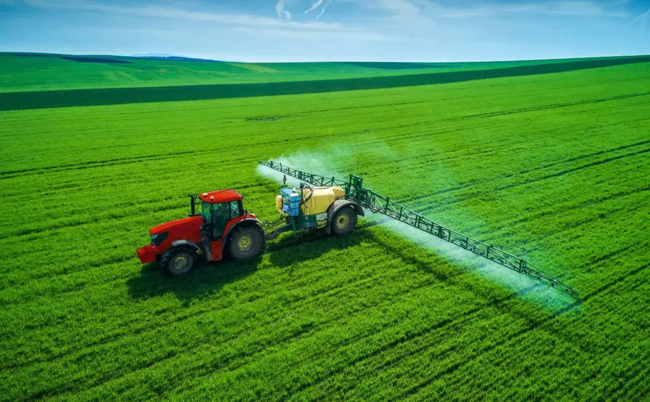 Czerwony traktor z opryskiwaczem na zielonym polu. Opryskiwacz rozpyla substancję, która może zawierać sól potasową, składnik odżywczy dla roślin.