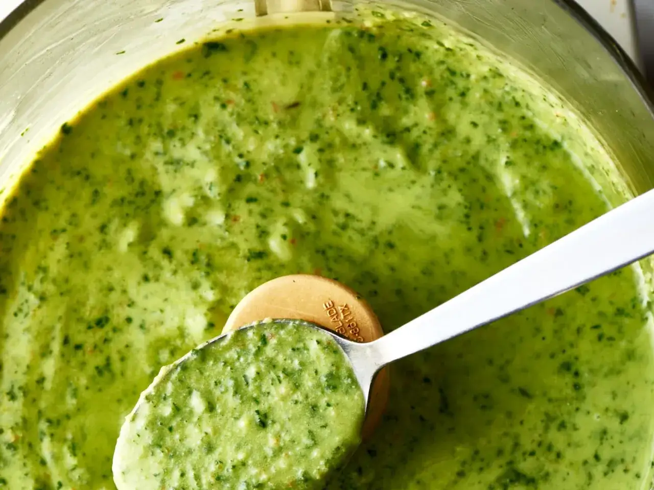 Cuchara sirviendo cremosa pasta con pesto, con trocitos de albahaca y queso.
