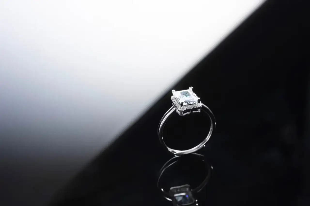 Elegancka fotografia biżuterii: srebrny pierścionek z prostokątnym, błyszczącym kamieniem otoczonym mniejszymi cyrkoniami, odbijający się w czarnej powierzchni.