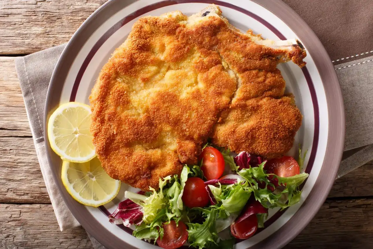 Cotoletta alla milanese dorada y crujiente, servida con rodajas de limón y una ensalada fresca de tomates y lechugas.