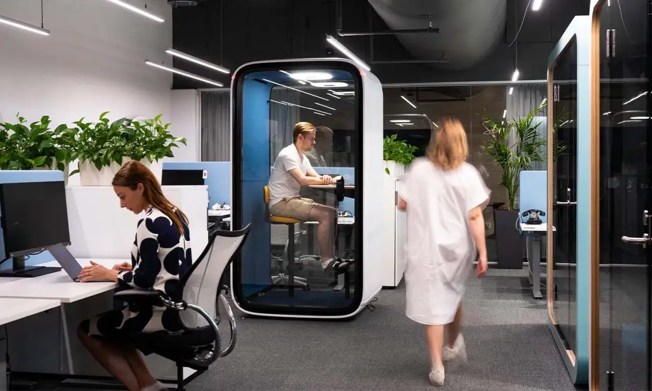 Dans ce bureau moderne, une femme travaille sur son ordinateur portable, tandis qu'un homme est dans une cabine insonoris&eacute;e pour &eacute;viter le bruit au travail. Une autre personne passe.