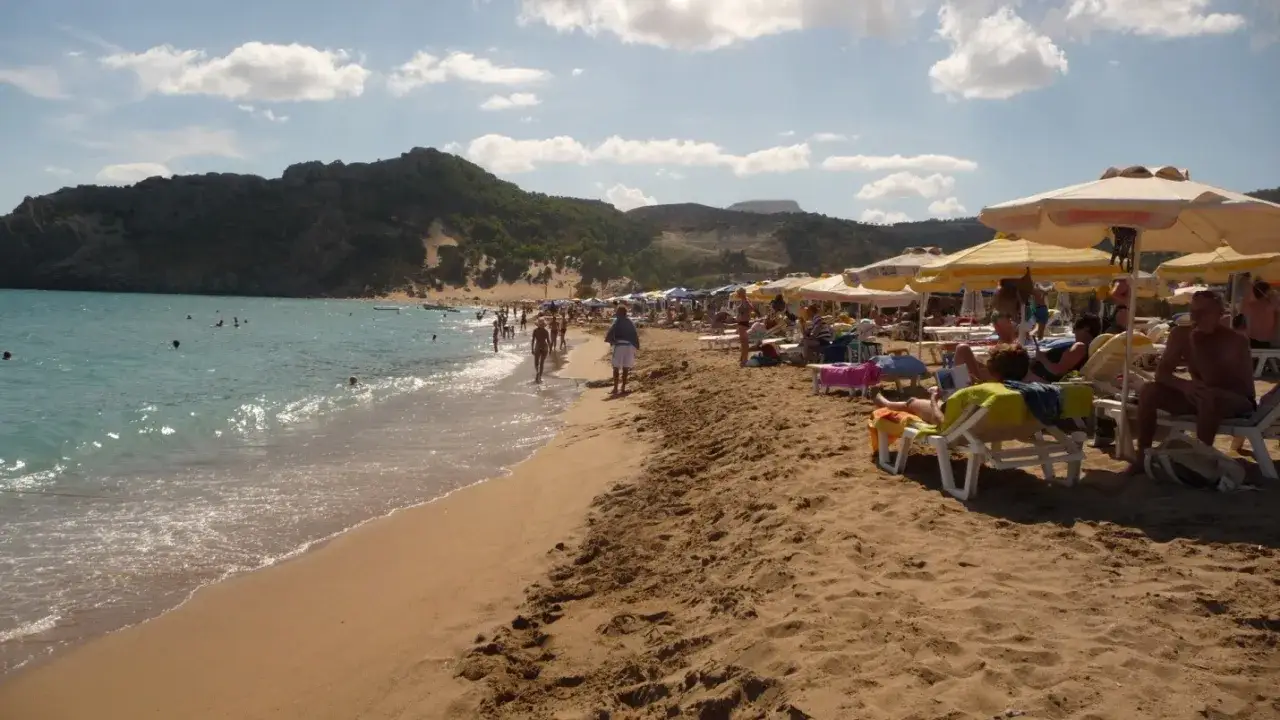 Słoneczny dzień na plaży Tsambika. Ludzie kąpią się w turkusowej wodzie, inni odpoczywają na leżakach pod parasolami.