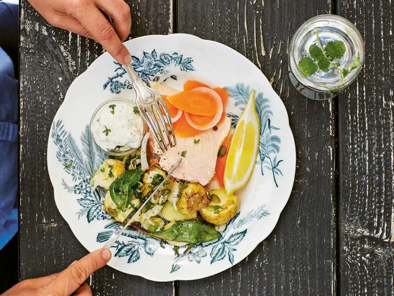 En tallrik med inkokt lax, potatis, gr&ouml;nsaker och en citronskiva. En person sk&auml;r i laxen med en kniv och gaffel.