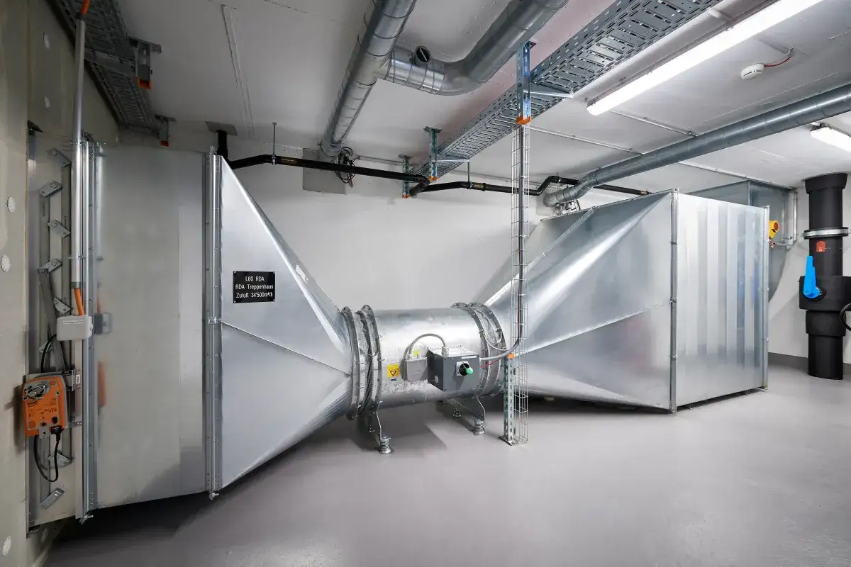 Große Lüftungsanlage für mechanische Entrauchung im Treppenhaus, mit auffälligen Metallkanälen und einem zentralen Ventilator.