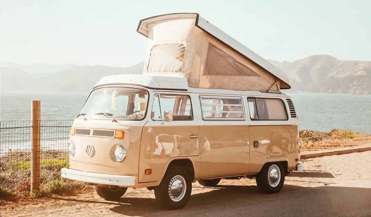 Un combi VW beige et blanc, toit relevé, stationné près de la mer, prêt pour une aventure.