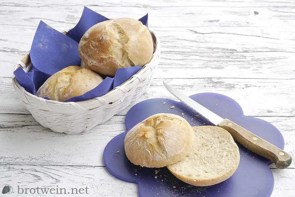 Frisch gebackenes Brot zu fest. Ein Korb mit Brötchen und ein aufgeschnittenes Brötchen auf einem lila Brett mit Messer.