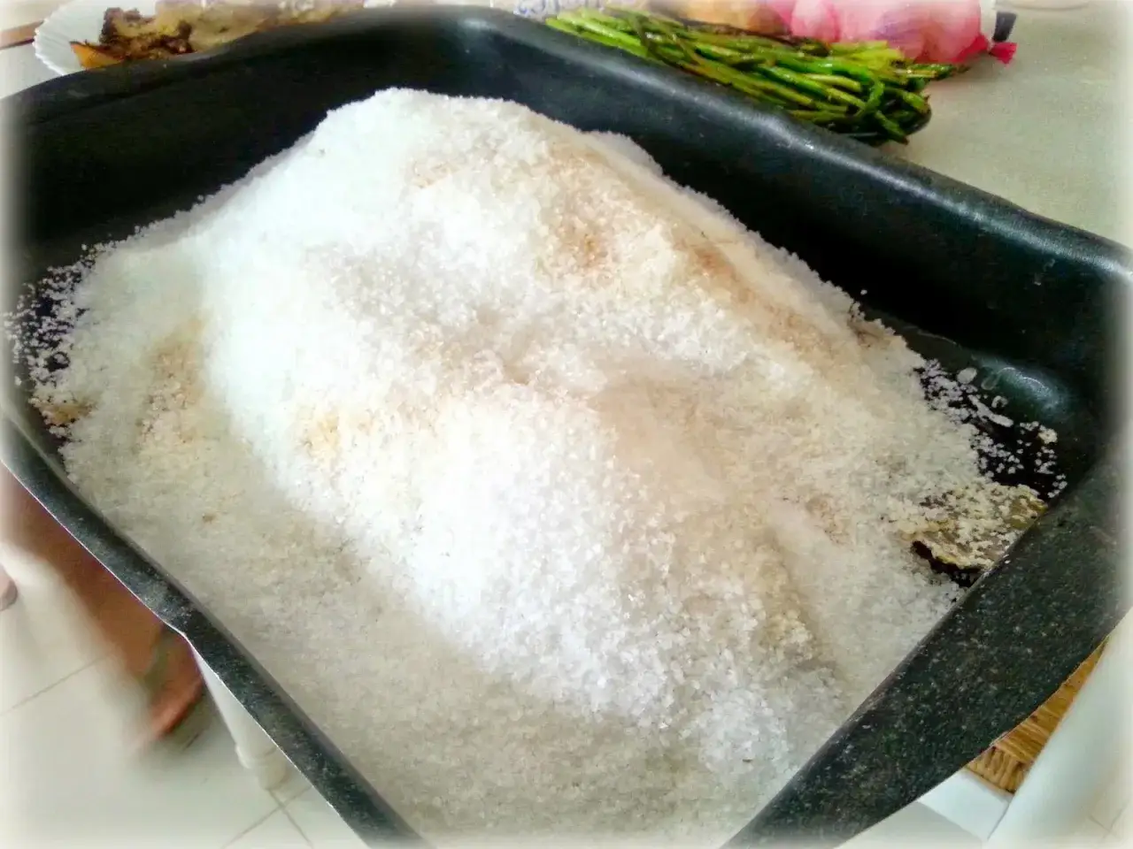 Dorada a la sal lista para hornear, cubierta con una gruesa capa de sal gruesa. En el fondo se aprecian espárragos y cebolla.