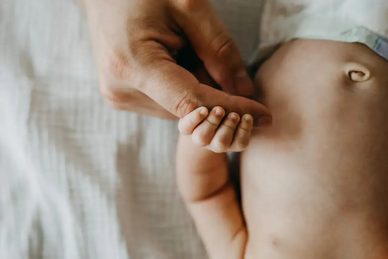 Dłoń rodzica obejmuje maleńką dłoń noworodka. Widoczny jest brzuszek dziecka z pępkiem, który może być oznaką, że zle gojacy sie pepek u noworodka wymaga uwagi.