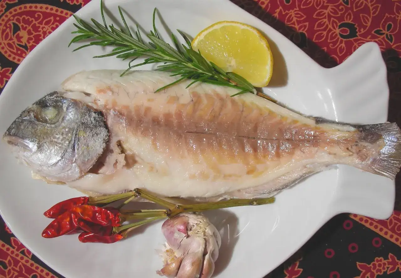 Dorada co to za ryba? Gotowana dorada z cytryną, rozmarynem, czosnkiem i papryczkami chili na białym talerzu.