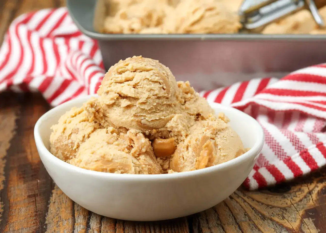 Schüssel mit cremigem erdnussbutter eis, garniert mit Karamellstücken. Im Hintergrund ein Blech mit mehr Eis und ein rot-weiß gestreiftes Tuch.