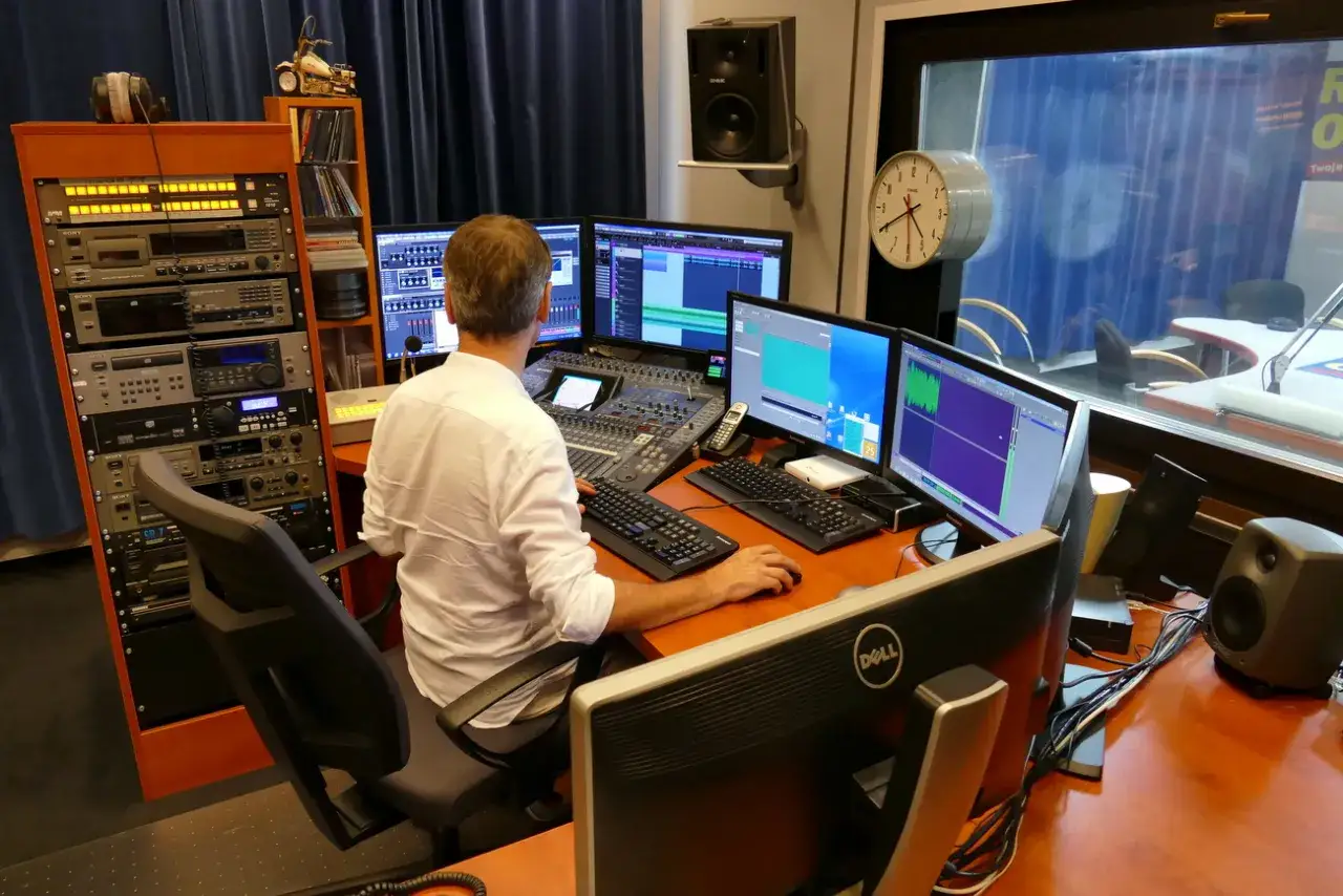 Mężczyzna w studiu nagraniowym, przy konsoli dźwiękowej i monitorach, przygotowuje audycję dla **radio park**.