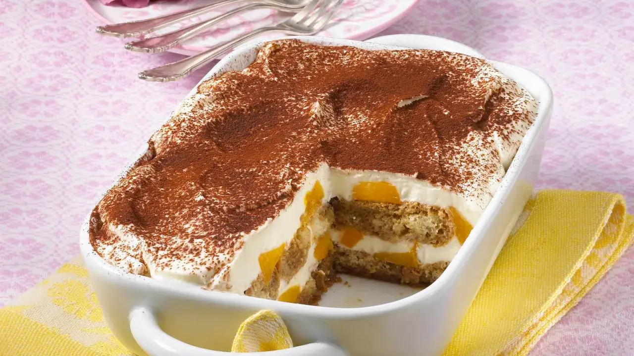 Ein cremiges Pfirsich Tiramisu, bestäubt mit Kakao, in einer weißen Auflaufform. Saftige Pfirsichstücke und Löffelbiskuits sind geschichtet.