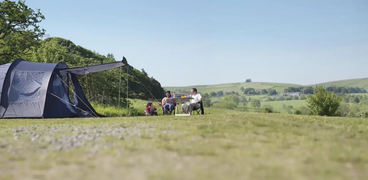 A family enjoys a sunny day camping alone. A blue tent is set up on a grassy hill, with a family sitting at a table nearby.