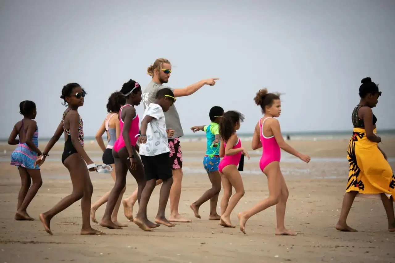 Un groupe d'enfants, guidés par un adulte, marche sur la plage. Une scène qui illustre le mouvement d'éducation populaire, favorisant l'apprentissage et le partage en plein air.