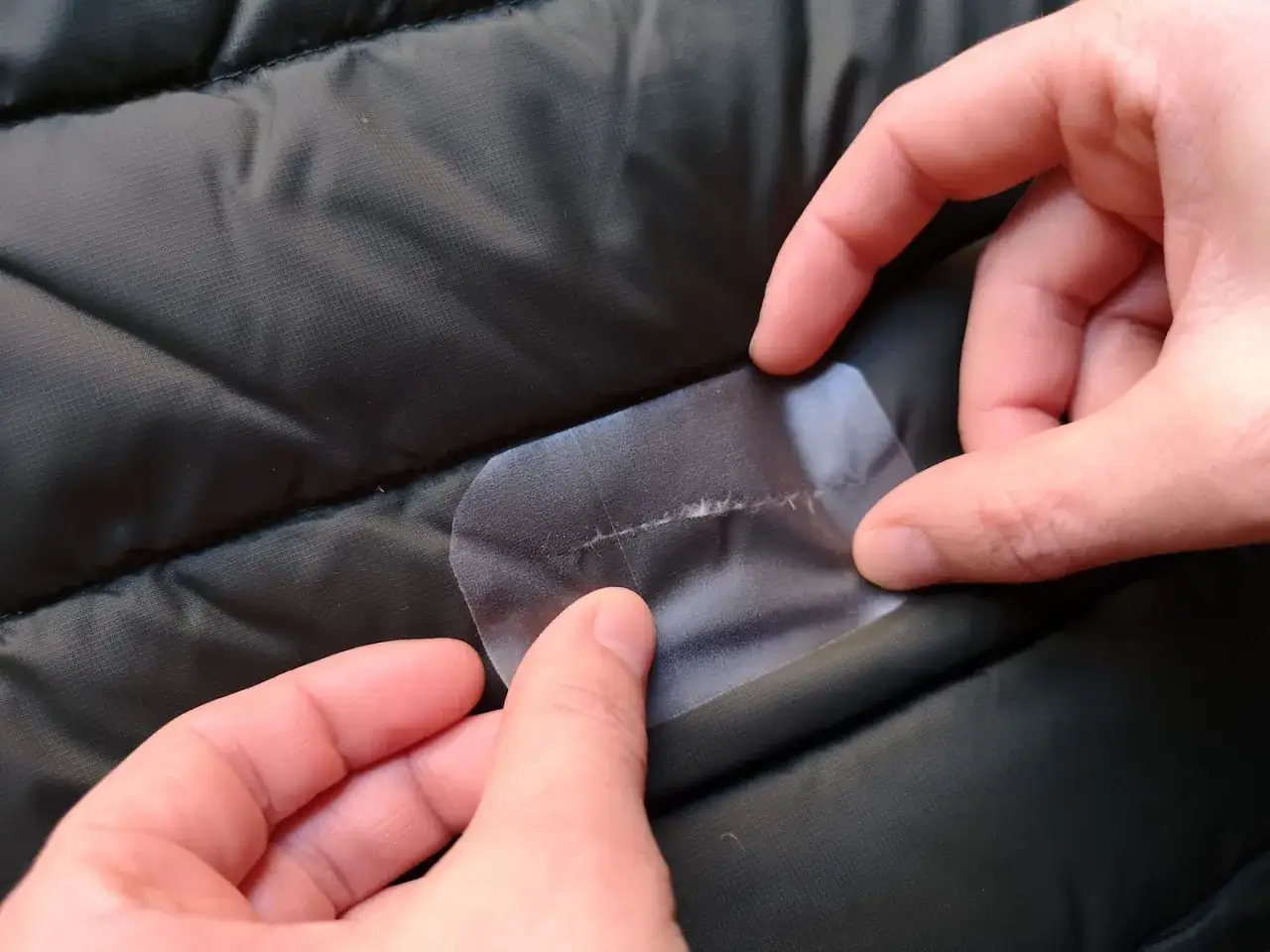 Hände flicken ein kleines Loch in einer Daunenjacke mit transparentem Klebeband.