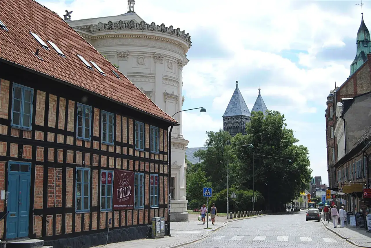 En korsvirkesbyggnad med blå fönster och ett rött tegeltak i förgrunden. I bakgrunden syns Lunds domkyrka och en vit, rund byggnad. En gata med kullersten leder bort mot domkyrkan. Kanske sveriges vackraste stad?