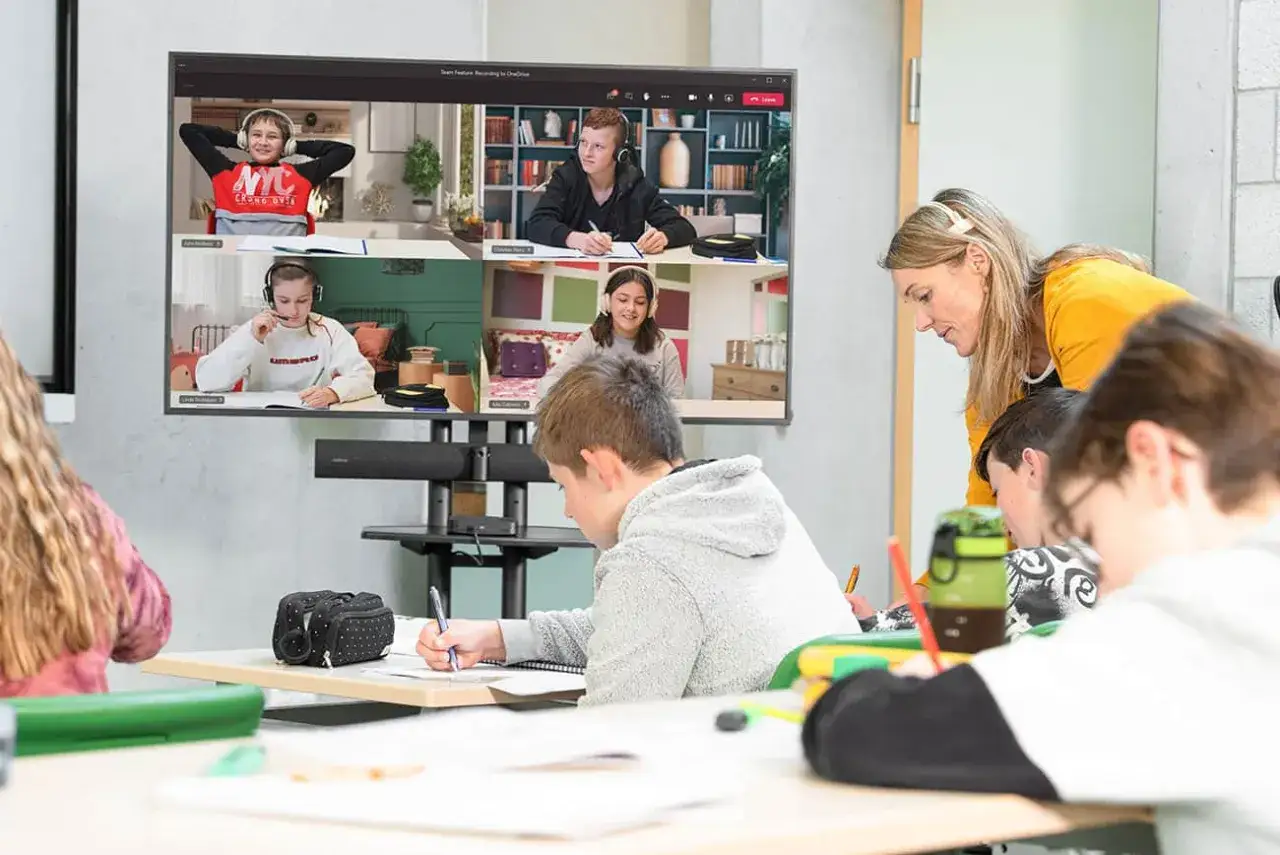 Lehrerin unterstützt Schüler bei der Projektarbeit Definition. Ein Bildschirm zeigt eine Videokonferenz mit anderen Schülern.
