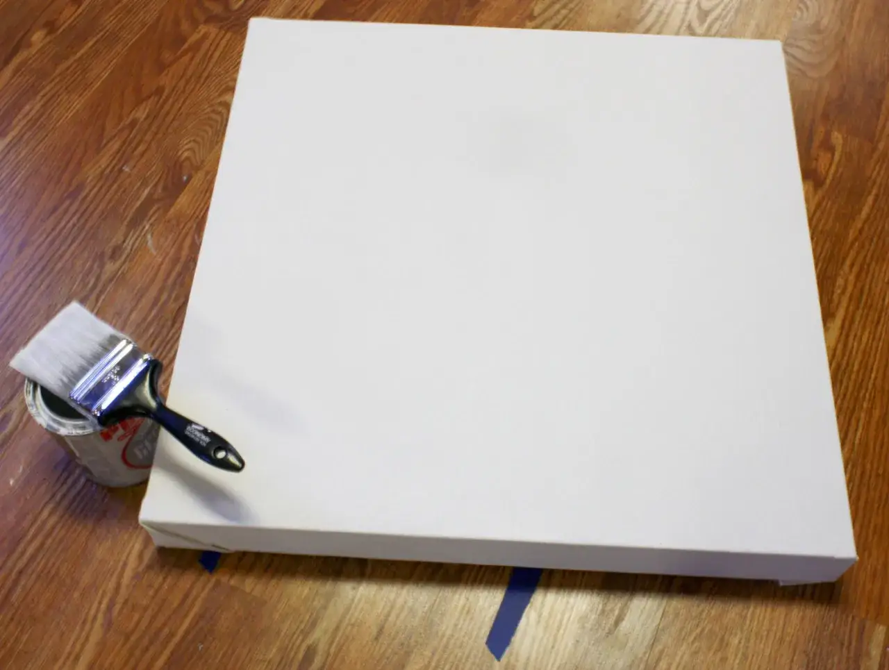 Un pinceau trempé dans une boîte de peinture est prêt à être utilisé sur un grand support de peinture acrylique blanc, posé sur un parquet en bois.