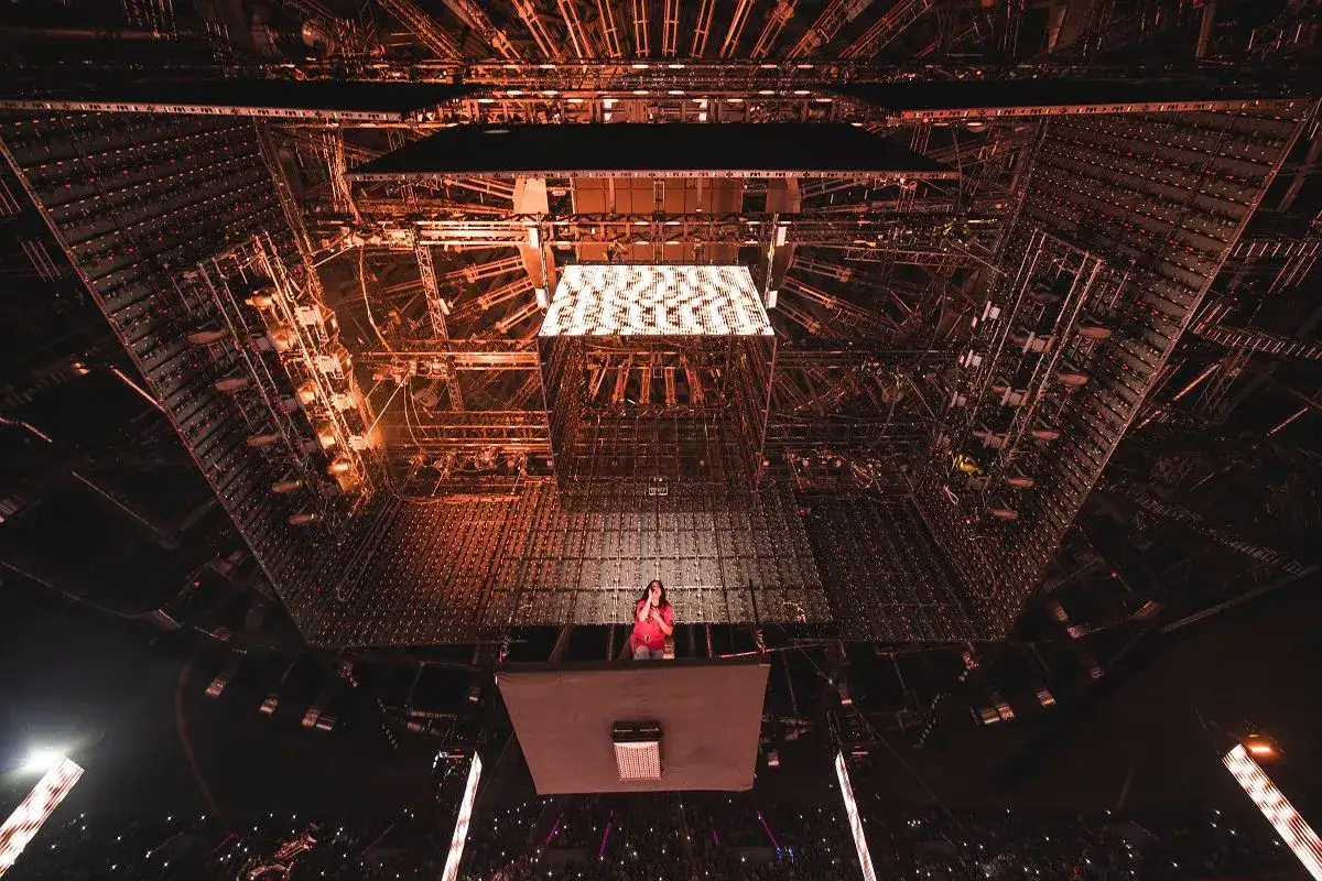 Billie Eilish na koncercie, unosząca się nad tłumem na platformie, otoczona skomplikowaną konstrukcją oświetleniową.