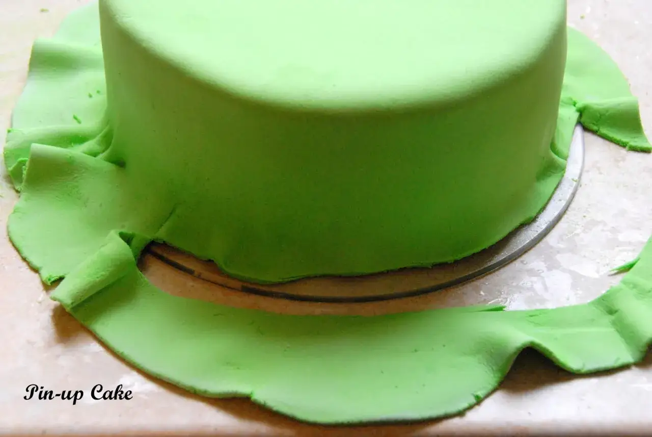 Zielony tort pokryty masą cukrową, kt&oacute;ra tworzy falbankę u dołu. To właśnie masa cukrowa, czyli fondant, co to jest i jak się ją stosuje.
