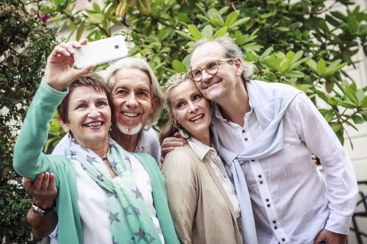 Vier lachende Freunde im Alter machen ein Selfie vor grünem Laub.
