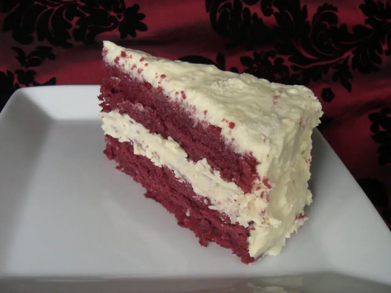 En bit saftig red velvet-t&aring;rta med kr&auml;mig frosting p&aring; en vit tallrik.