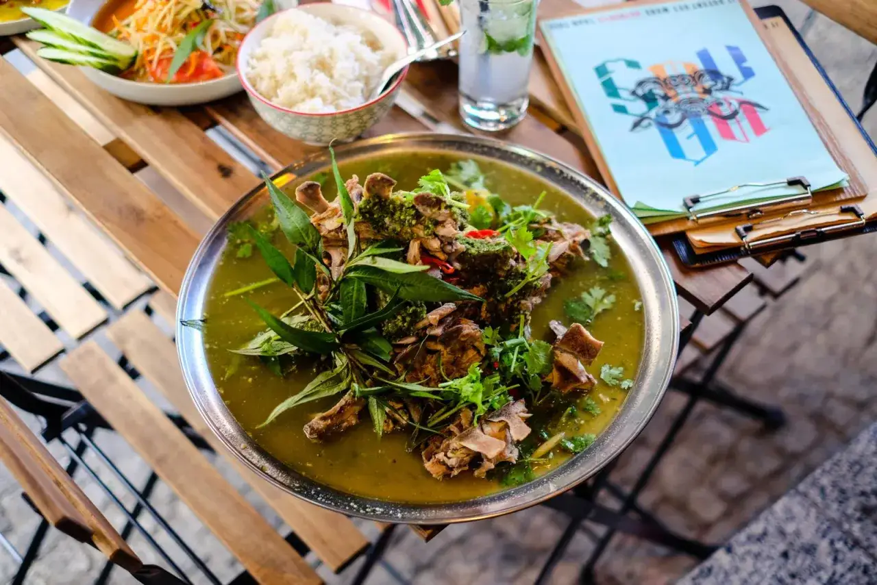 Ein herzhaftes Gericht mit Fleisch und Kr&auml;utern in einer gr&uuml;nen Br&uuml;he, serviert auf einem Tisch in einem Thai Restaurant Berlin. Daneben Reis und ein Drink.