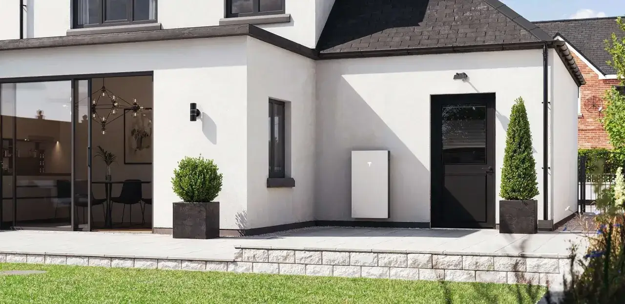 Un moderno Tesla Powerwall blanco est&aacute; montado en la pared exterior de una casa blanca, junto a una puerta negra y un jard&iacute;n verde.