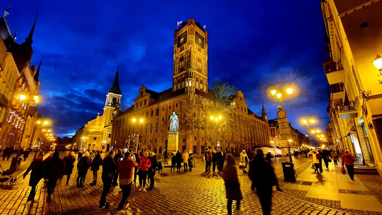 Wieczorne atrakcje Torunia: Rynek Staromiejski z Ratuszem i pomnikiem Kopernika, oświetlony brukowany deptak pełen spacerowicz&oacute;w.