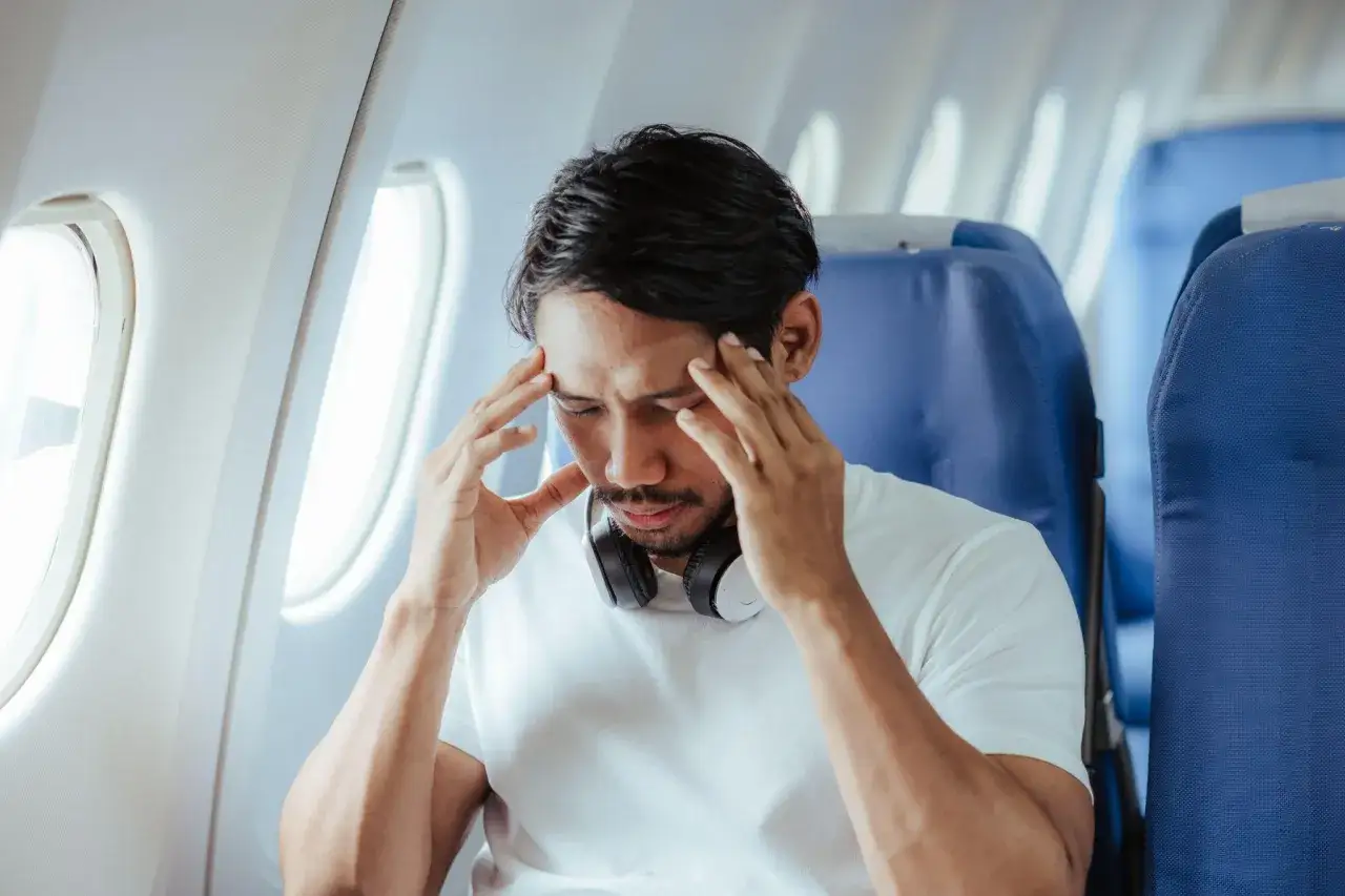 Man med huvudvärk i flygplansstol, verkar sjuk efter utlandsresa. Huvudtelefoner runt halsen.