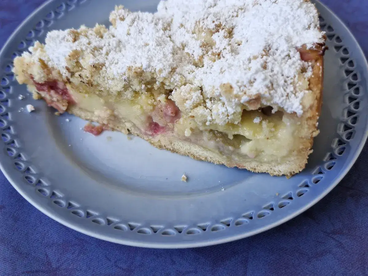 Ein St&uuml;ck Rhabarberkuchen mit Streuseln, best&auml;ubt mit Puderzucker, auf einem blauen Teller.