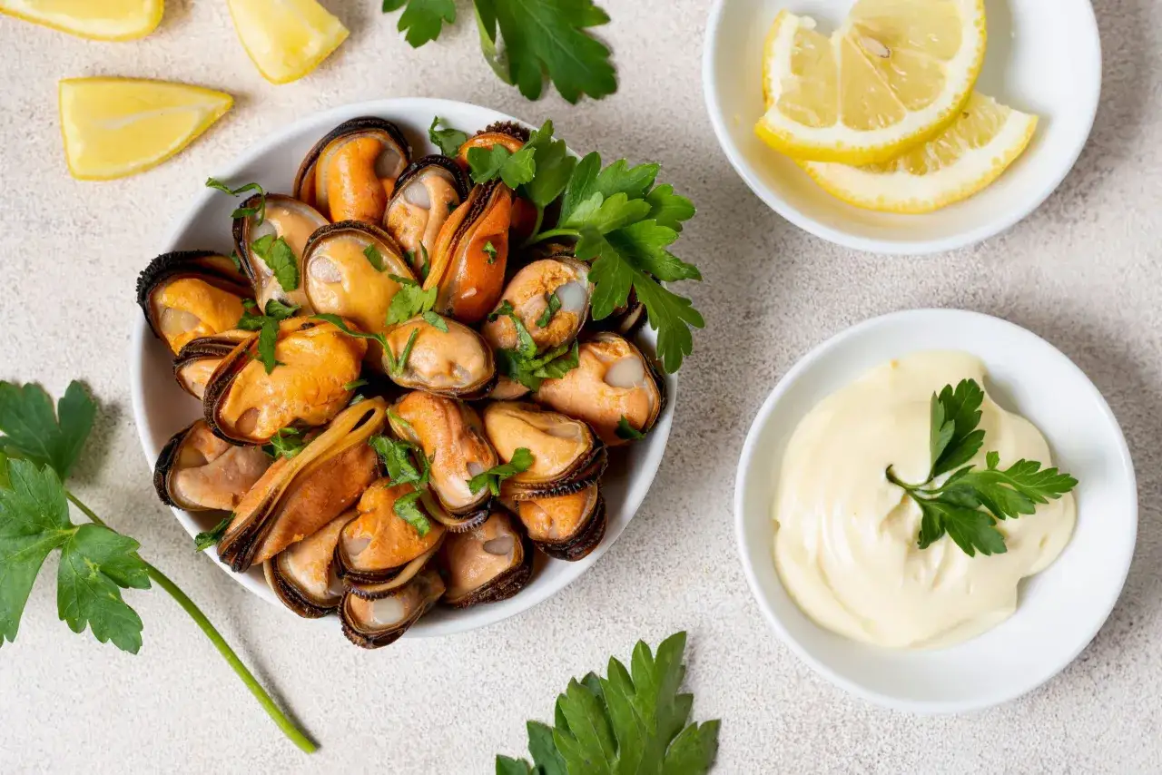 Un plato lleno de mejillones cocidos, adornados con perejil fresco. A su lado, gajos de limón y un cuenco con mayonesa, para disfrutar de qué son los mejillones.