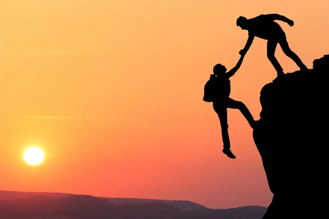 Silhouettes of two hikers reaching for each other on a cliff at sunset. Prawdziwy autorytet to ten, kto podaje pomocną dłoń.