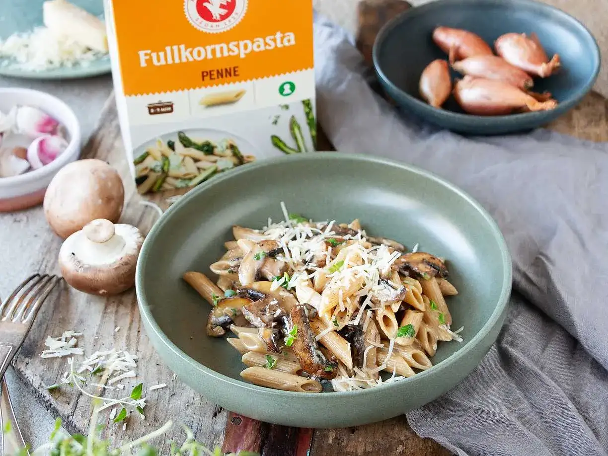 En skål med krämig svamp pasta, toppad med riven ost och färska örter. Bredvid syns en förpackning fullkornspasta penne, champinjoner och schalottenlök.
