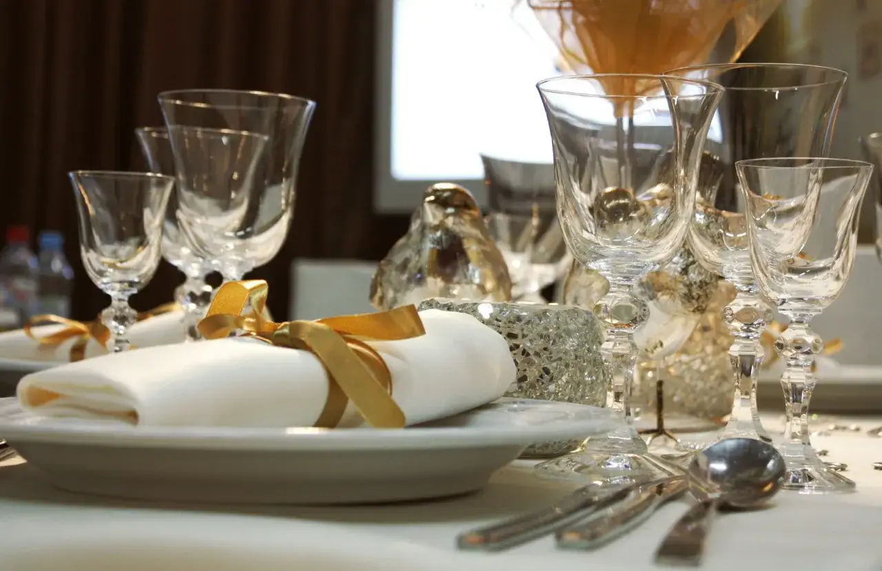 Une table dressée avec élégance, verres en cristal, serviettes blanches ornées de rubans dorés, et des décorations scintillantes. Un bel exemple des arts de la table.