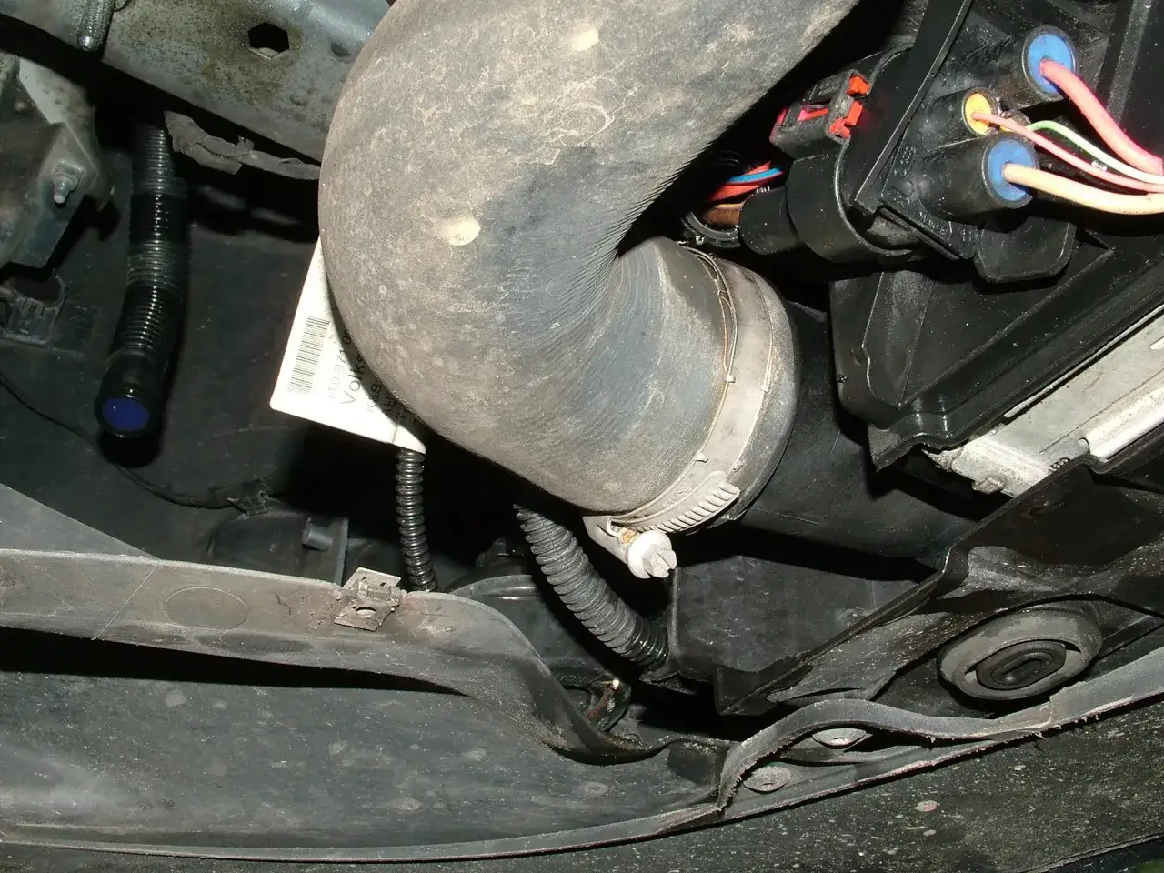 A car's engine bay shows a large hose, electrical connectors, and a drip of coolant on a black corrugated tube, indicating a car leaking coolant.