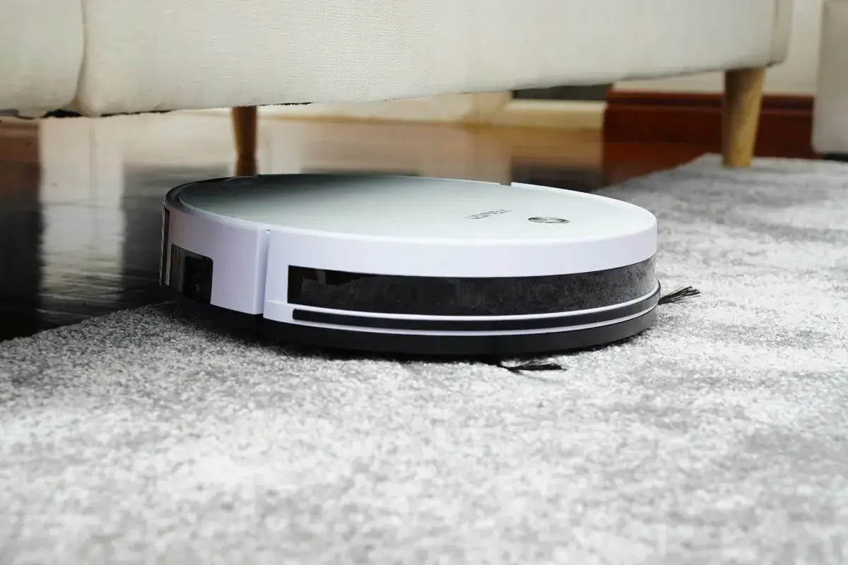 Robotdammsugare navigerar över en grå, luddig ullmatta under en soffa.