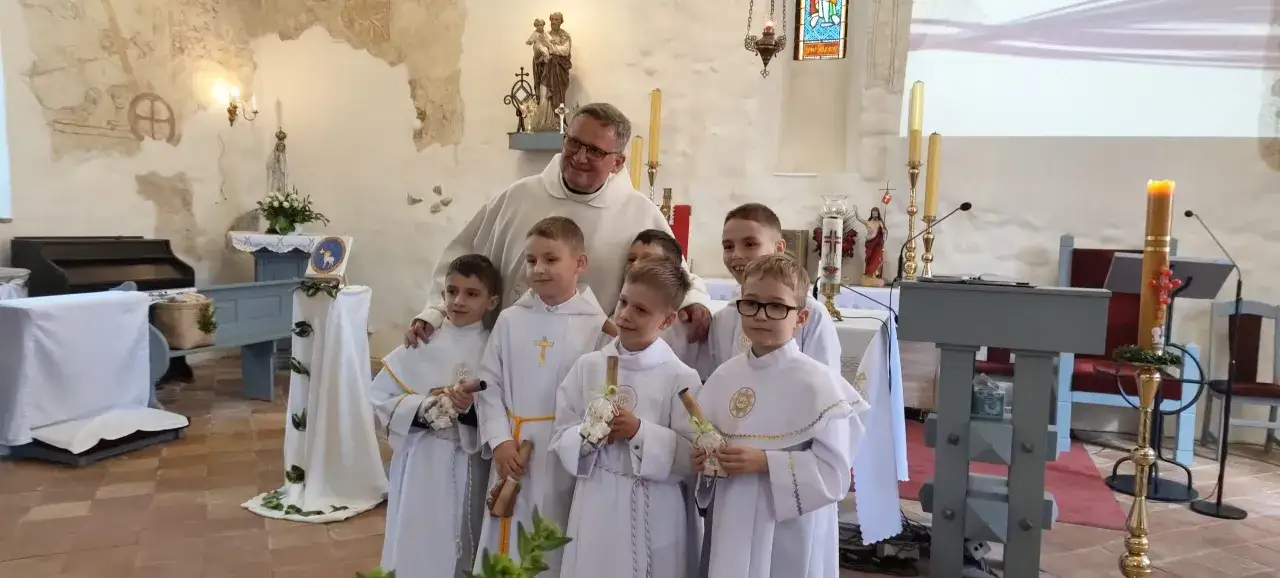 Ksiądz z uśmiechem pozuje do zdjęcia z chłopcami w wieku komunijnym, ubrani w białe alby.