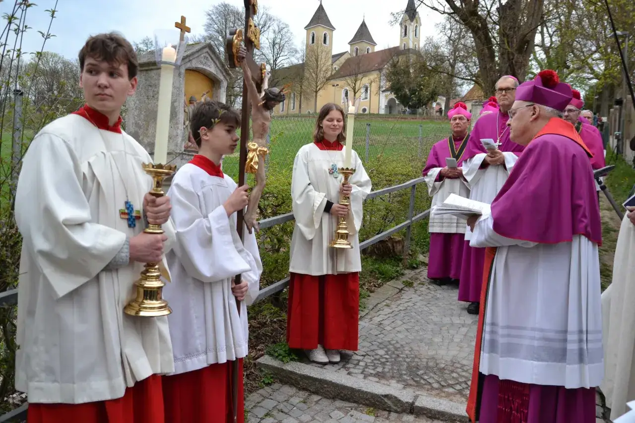 Jugendliche tragen Kerzen und ein Kreuz, w&auml;hrend Bisch&ouml;fe in lila Gew&auml;ndern bei den Kreuzweg Stationen vor einer Kirche stehen.