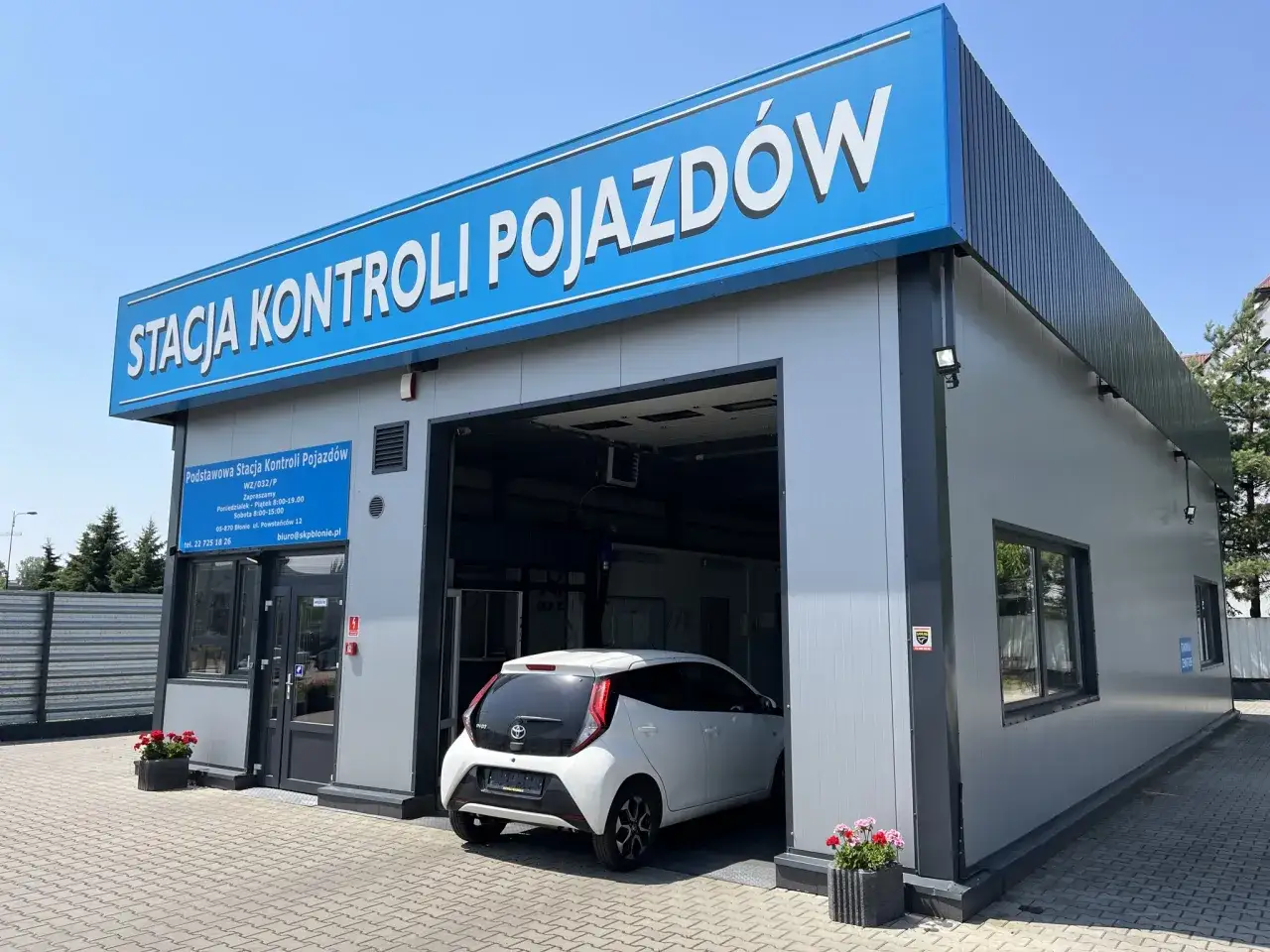 Stacja kontroli pojazdów z białym samochodem w środku. Dowiedz się, ile teraz kosztuje przegląd samochodu, sprawdzając godziny otwarcia i dane kontaktowe stacji.