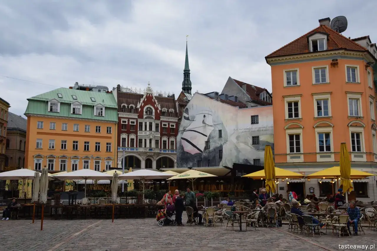Ryga co zobaczyć: kolorowe kamienice, mural z ptakiem i kawiarniane ogródki na brukowanym placu.