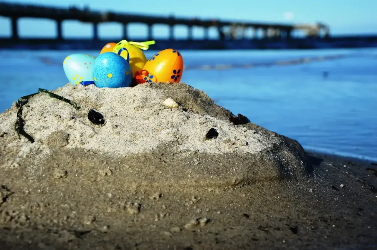 Bunte Ostereier auf einer Sanddüne am Meer. Ein Pier im Hintergrund. Perfekt für Veranstaltungen an Ostern am Strand.
